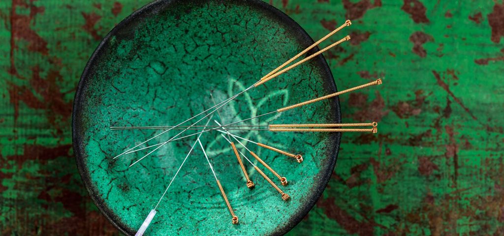 Close-up of acupuncture needles arranged in a decorative green ceramic bowl on a textured green background, symbolizing traditional healing practices.