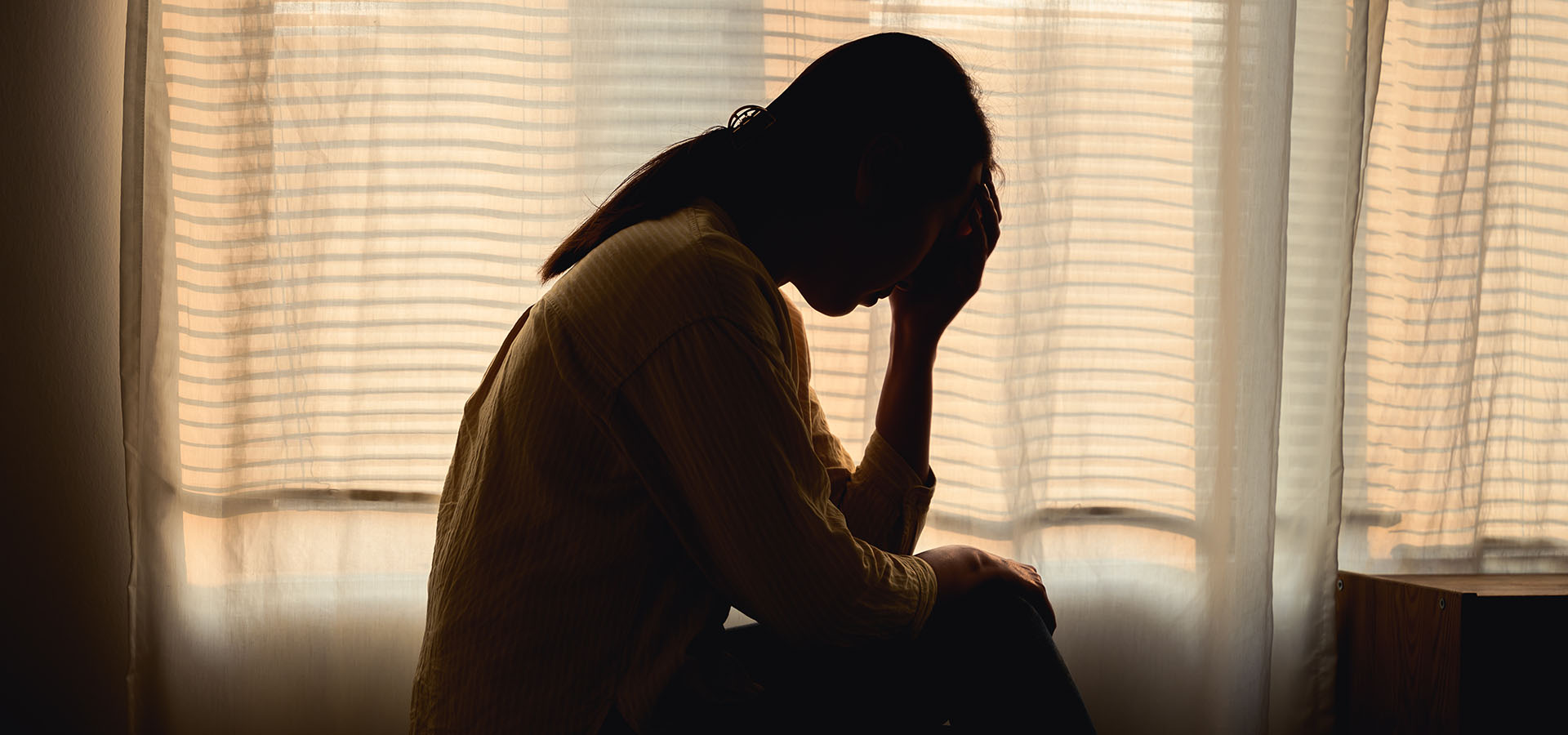 Silhouette of a person sitting with their head in their hand in front of a window with soft light filtering through, depicting stress, sadness, or emotional struggle.