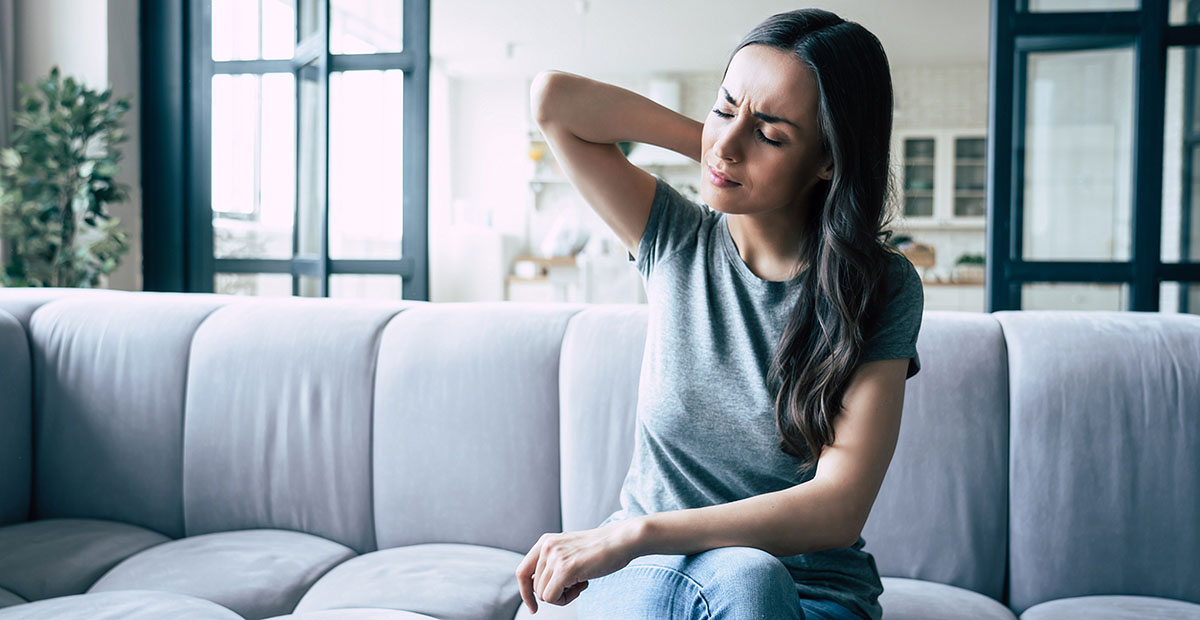 Woman sitting on a sofa holding her neck and shoulder in discomfort, illustrating pain relief or muscle tension.