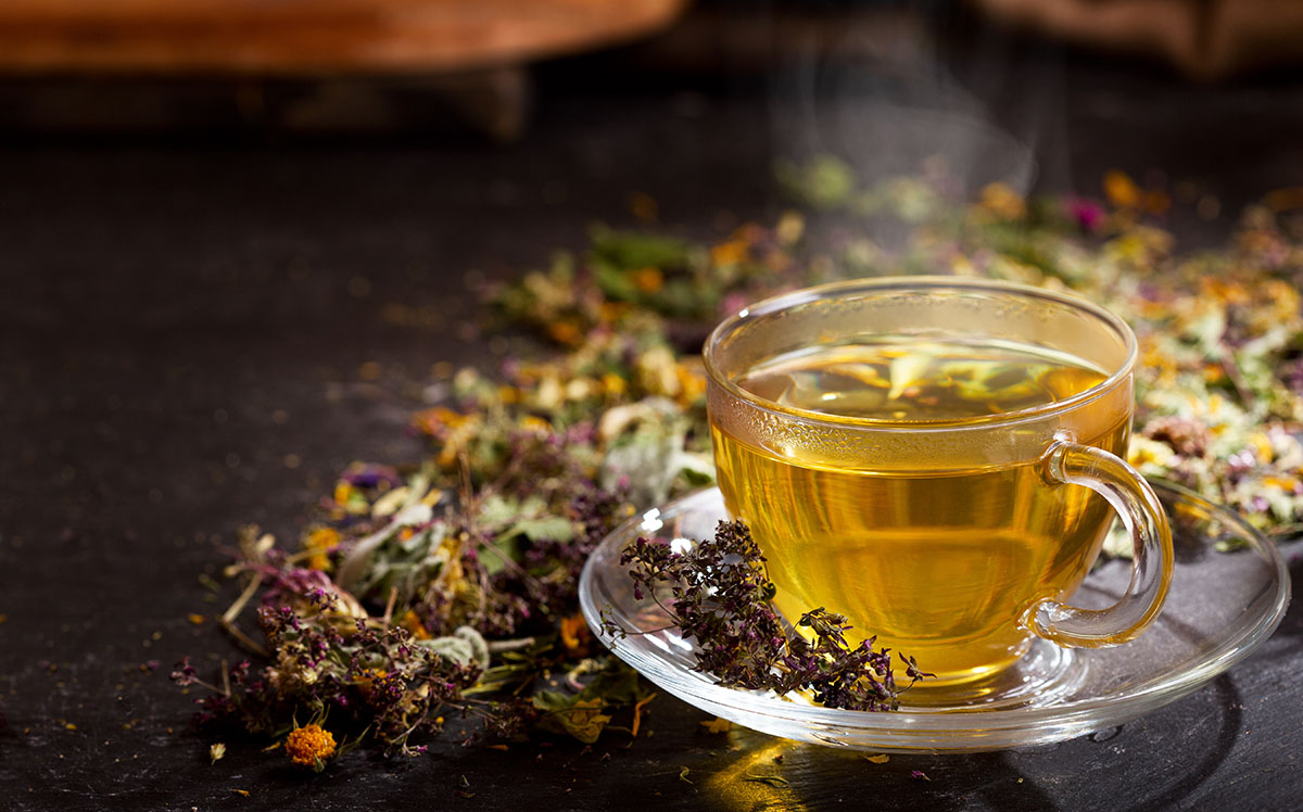 Glass cup of steaming herbal tea placed on a saucer surrounded by dried herbs and flowers, symbolizing natural remedies and relaxation.