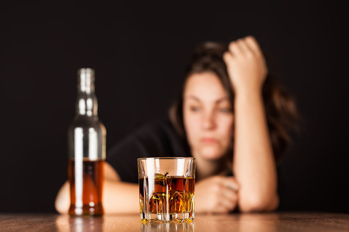 A glass of whiskey and a bottle of alcohol on a wooden table, with a blurred woman in the background appearing stressed or upset, symbolizing struggles with alcohol.