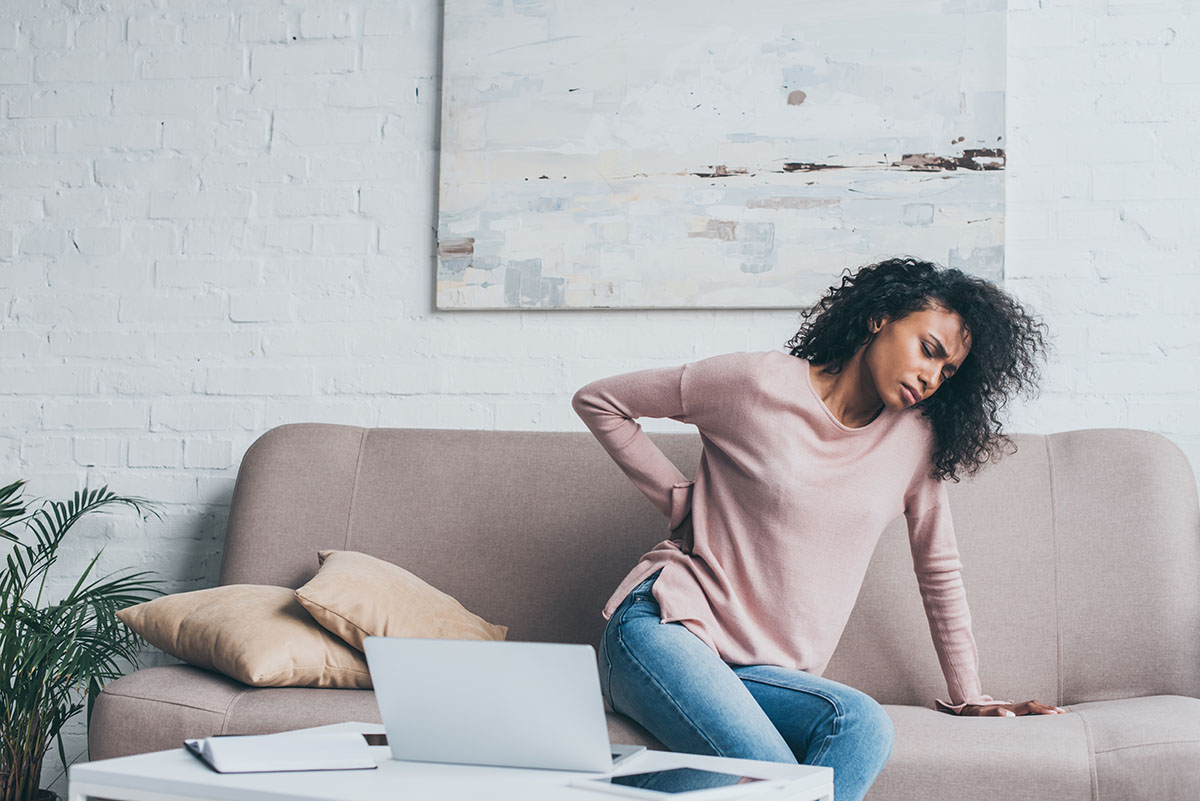 Young woman sitting on a couch holding her lower back in discomfort, highlighting the challenges of back pain and the importance of proper posture and physical care.