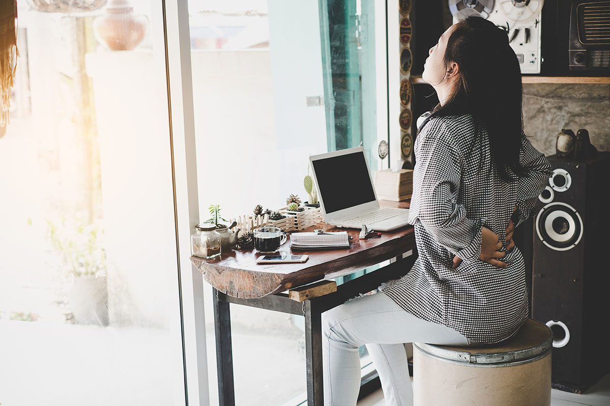 Young woman stretching her back at a home office desk to alleviate back pain, emphasizing the importance of proper posture and ergonomic workspace setup during remote work.