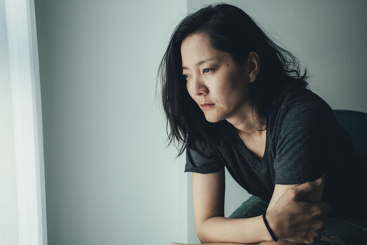 Close-up of a woman with long dark hair, sitting near a window, looking distressed and lost in thought, depicting feelings of anxiety, depression, or fatigue.