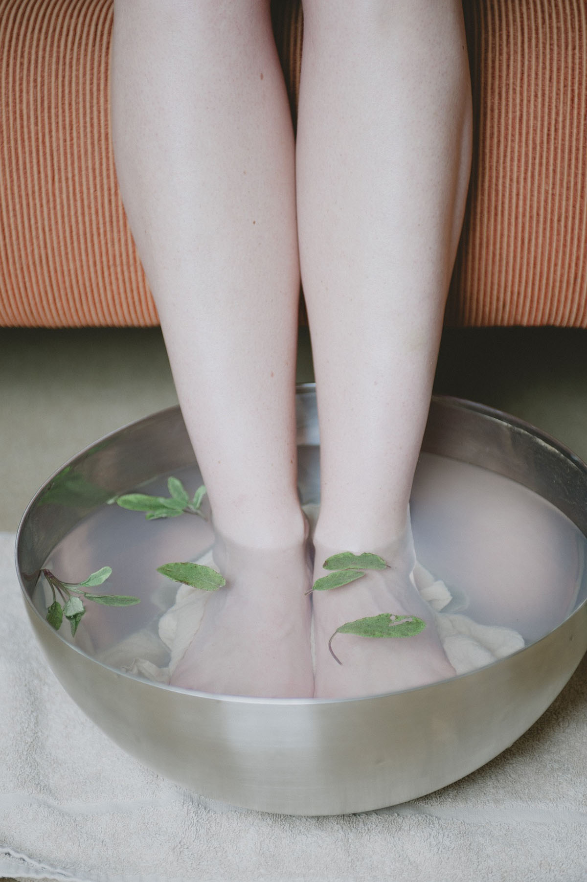 A person soaking their feet in a large metal bowl filled with water, epsom salts, and floating green leaves, promoting relaxation and natural foot care.