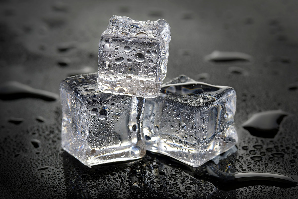 Close-up of three crystal-clear ice cubes stacked on a dark, wet surface with visible water droplets, symbolizing refreshment, cooling, and hydration.