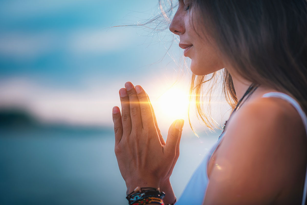 Woman in serene meditation at sunset, hands in prayer position with sunlight reflecting through, symbolizing mindfulness and inner peace.