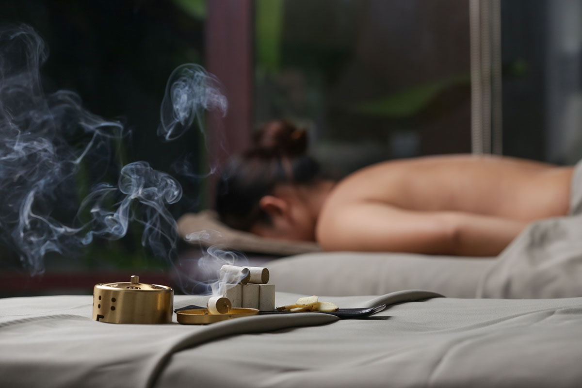 Relaxing spa scene with a woman lying on her stomach for therapy, accompanied by smoking moxa sticks and traditional tools in the foreground, emphasizing holistic wellness and healing.