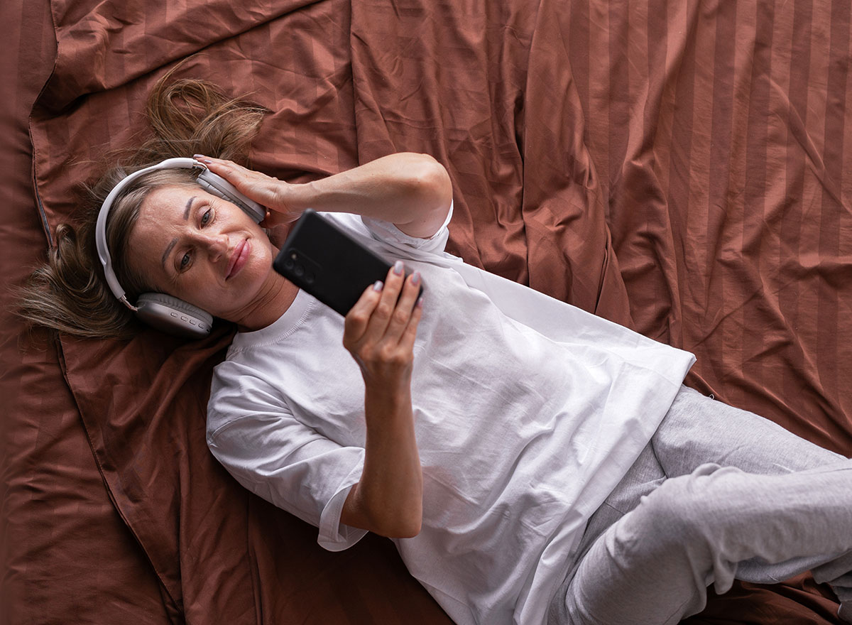 Middle-aged woman relaxing on a bed while listening to music through headphones and holding a smartphone, showcasing self-care and stress relief through technology and relaxation.