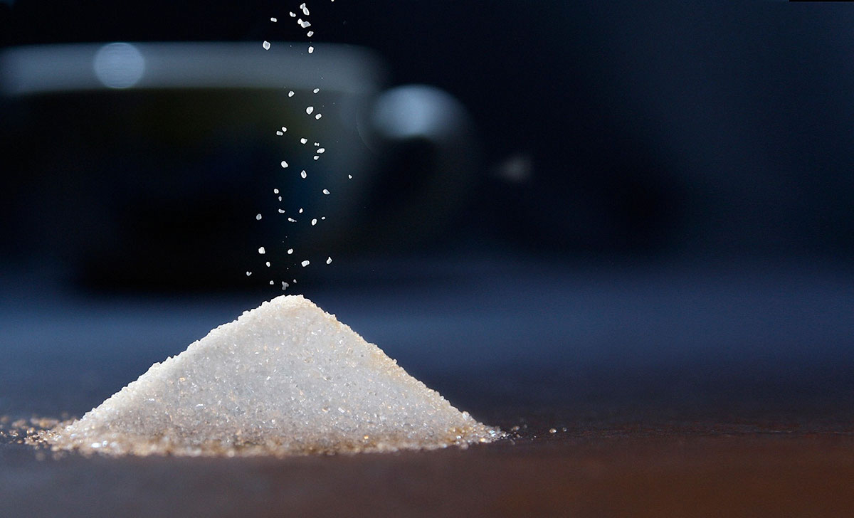 A pile of white granulated sugar on a dark surface with sugar crystals falling from above, highlighting the texture and detail of the granules.