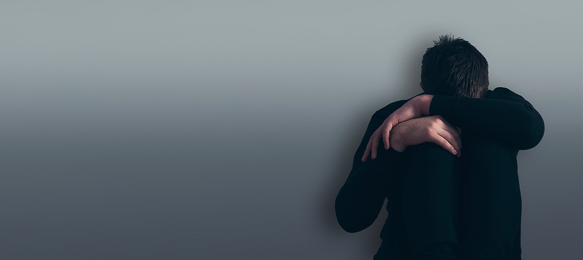 Man sitting on the floor with knees hugged, symbolizing loneliness and depression, against a minimalist gray background.