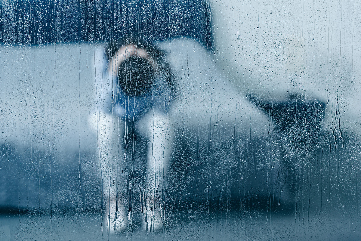 Depressed woman sitting on bed and holding head in hands through window with raindrops.