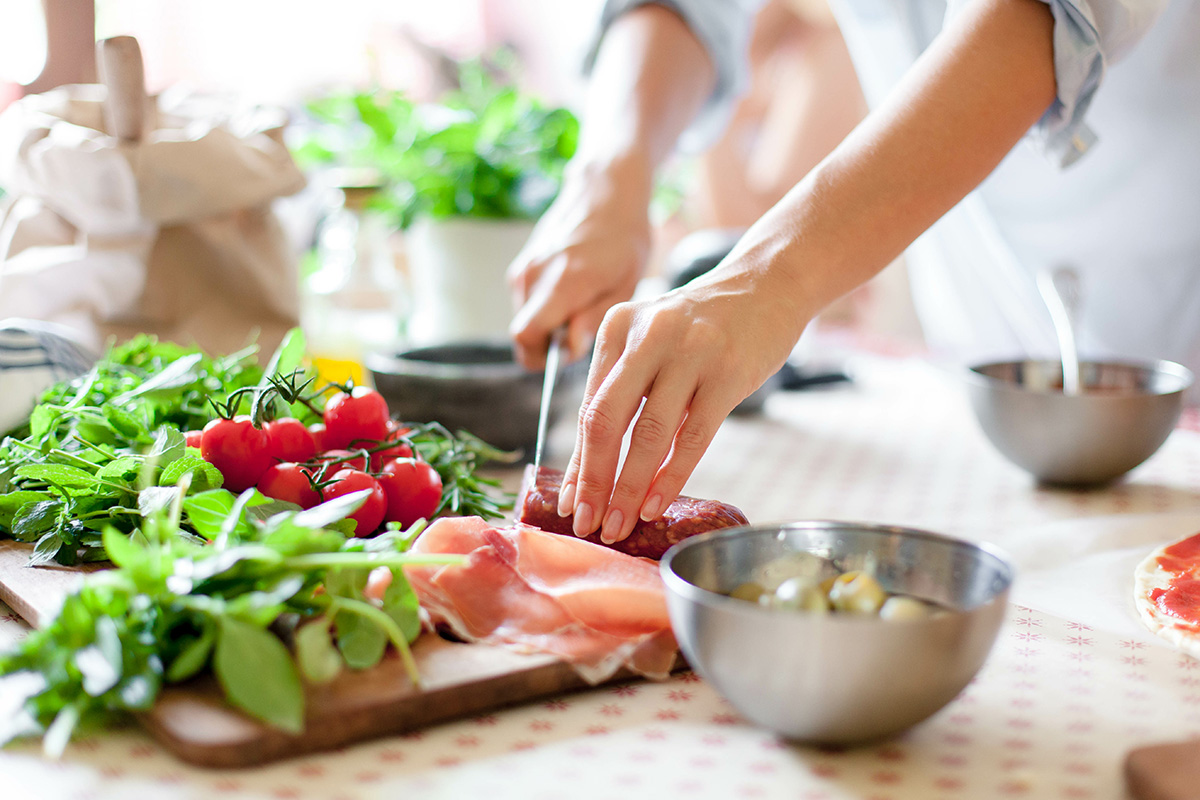 Woman is cooking in home kitchen. Female hands cut salami, vegetables, greens, tomatoes on table on wooden boards.