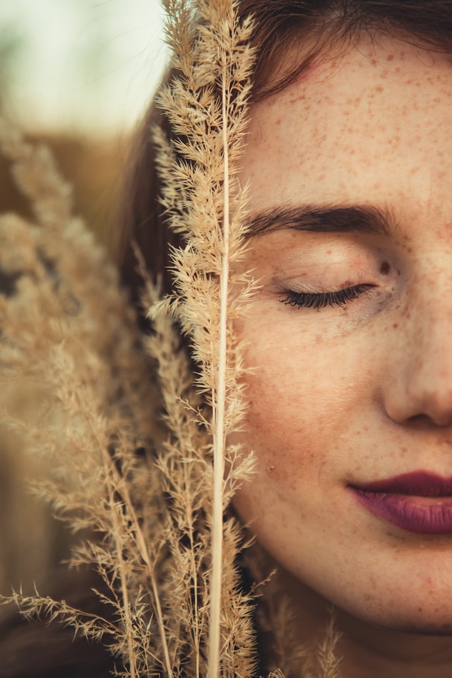 Close-up of a serene woman with freckles, eyes closed, surrounded by golden wheat in natural sunlight.