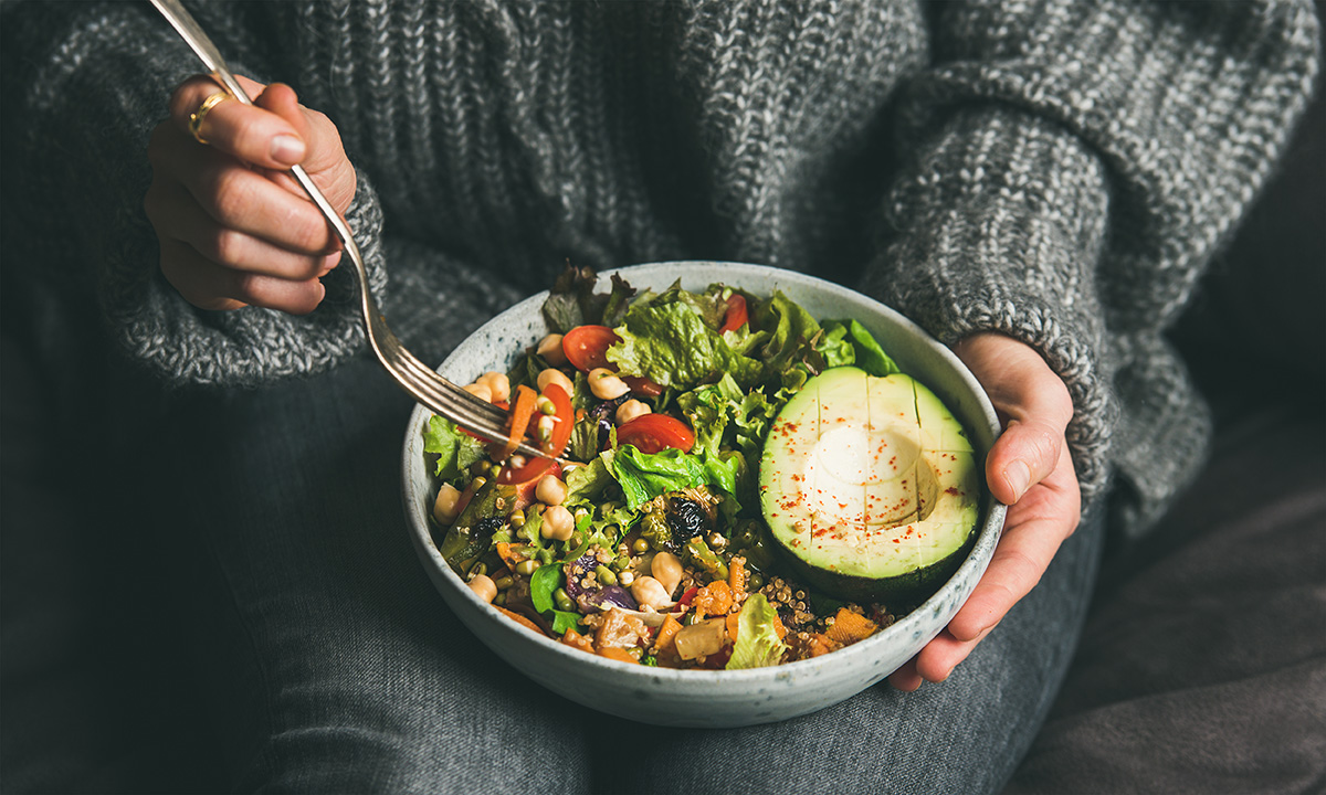 Woman in jeans and warm sweater holding bowl with fresh salad, avocado, grains, beans, roasted vegetables.