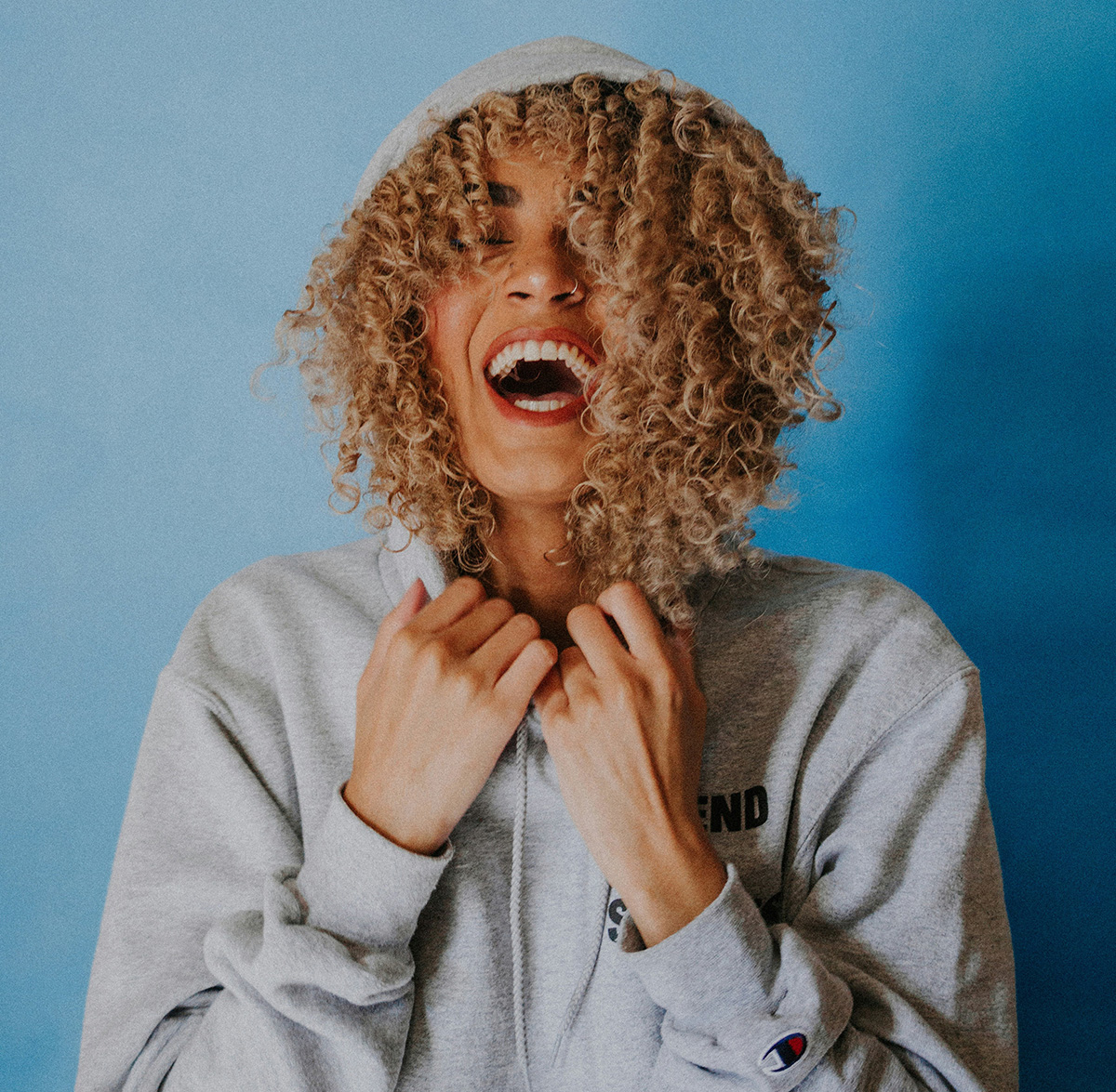 Joyful young woman with curly hair wearing a hoodie, laughing against a vibrant blue background.