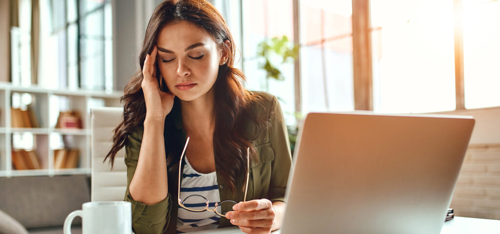 Tired business woman in stress works at a laptop while sitting at a table at home and holds her hand on her temples.