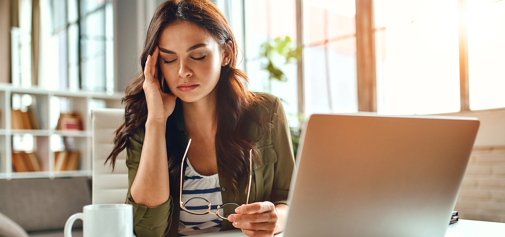 Tired business woman in stress works at a laptop while sitting at a table at home and holds her hand on her temples.