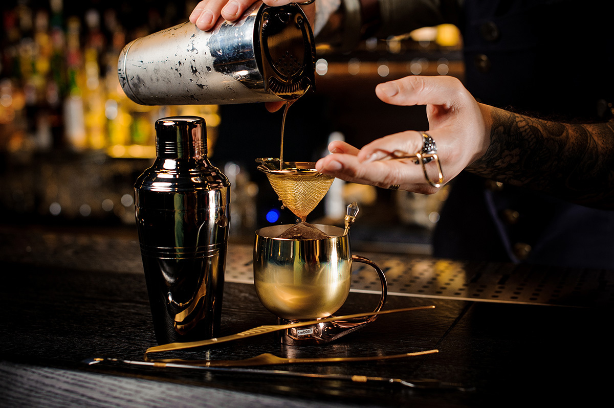 Bartender with tattoo pouring fresh and tasty summer drink from shaker into the vintage copper mug on bar counter